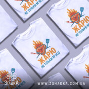 Футболка жарю м`ясо, біла, зображена з фронтального ракурсу, фон світло-сірий, принт з зображенням шматка м'яса з вогнем і кухонними приладами, текст "ЖАРЮ НЕ ТІЛЬКИ М'ЯСО" яскравими кольорами.