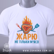 Футболка жарю м`ясо, біла, зображена з верхнього ракурсу, фон світло-сірий, принт з зображенням шматка м'яса з вогнем і кухонними приладами, текст "ЖАРЮ НЕ ТІЛЬКИ М'ЯСО" яскравими кольорами, на футболці є етикетка.
