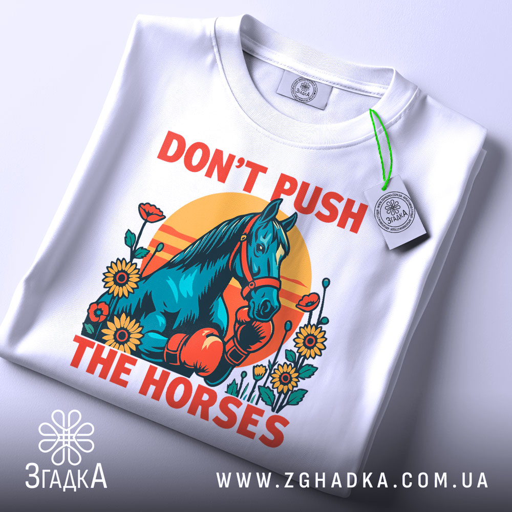 Футболка жарт і стиль. Біла футболка з принтом, на якому зображений кінь у рукавичках, оточений квітами на фоні сонця. Футболка складена на столі, з прикріпленою етикеткою, фон світлий, з легким градієнтом.