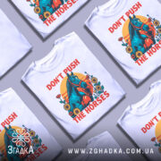 Футболка жарт і стиль. Біла футболка з яскравим принтом, на якому зображений кінь у рукавичках, оточений квітами на фоні жовто-оранжевого сонця. Футболка представлена на манекені з металевим блиском, фон нейтральний сірий.