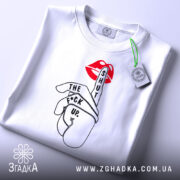 Футболка жест тиші, біла, складена на столі, фон світло-сірий, принт з червоними губами та рукою, що показує жест тиші, текст чорного кольору, на комірці видна етикетка.