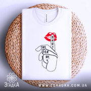 Футболка жест тиші, біла, складена на плетеній кошику, фон світло-сірий, принт з червоними губами та рукою, що показує жест тиші, текст чорного кольору.