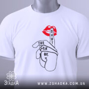 Футболка жест тиші, біла, складена на столі, фон світло-сірий, принт з червоними губами та рукою, що показує жест тиші, текст чорного кольору, на комірці видна етикетка.