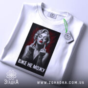 Футболка жінка тату білого кольору, зображена в горизонтальному ракурсі, фон світлий, принт з чорно-білою жінкою з червоними губами та татуюваннями, текст "ВЖЕ НЕ МОЖУ" під зображенням, на футболці є етикетка з зеленим шнурком.