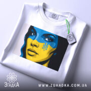 Футболка жінка Україна, біла, зображена на столі, фон світлий, принт з обличчям жінки з синьо-жовтим малюнком, на футболці є етикетка з зеленим шнурком.