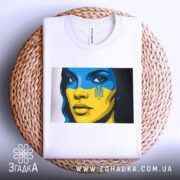 Футболка жінка Україна, біла, зображена на плетеній кошику, фон світлий, принт з обличчям жінки з синьо-жовтим малюнком, футболка акуратно складена.