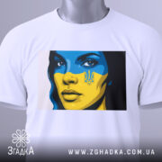 Футболка жінка Україна, біла, зображена на столі, фон світлий, принт з обличчям жінки з синьо-жовтим малюнком, на футболці є етикетка з зеленим шнурком.