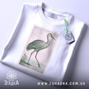 Футболка журавель стиль, біла футболка, складена на столі, з принтом журавля, фон світло-сірий, на футболці є етикетка з логотипом.