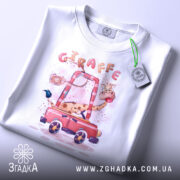 Футболка жираф у машині, біла, лежить на столі, фон світло-сірий, принт з жирафом у червоній машині, з етикеткою на рукаві.
