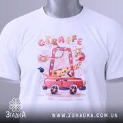 Футболка жираф у машині, біла, лежить на столі, фон світло-сірий, принт з жирафом у червоній машині, з етикеткою на рукаві.
