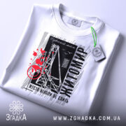 Футболка Житомир місто моє, біла футболка з коротким рукавом, розкладена на поверхні, фон світлий, принт містить чорні та червоні елементи з написом 'Житомир' і малюнком, що нагадує міст, на футболці є зелений ярлик.