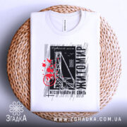 Футболка Житомир місто моє, біла футболка з коротким рукавом, розкладена на плетеній кошику, фон світлий, принт містить чорні та червоні елементи з написом 'Житомир' і малюнком, що нагадує міст.