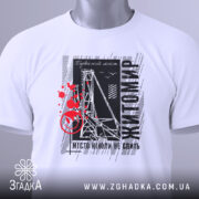 Футболка Житомир місто моє, біла футболка з коротким рукавом, розкладена на поверхні, фон світлий, принт містить чорні та червоні елементи з написом 'Житомир' і малюнком, що нагадує міст, на футболці є зелений ярлик.