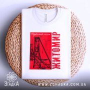 Футболка Житомир, біла, складена на плетеній кошику, фон світло-сірий, принт червоний з малюнком підвісного мосту та написом 'Житомир', видно етикетку на комірці.