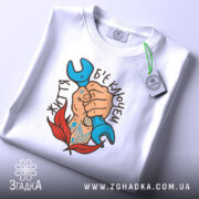 Футболка життя б`є ключем. Біла футболка складена, на ній видно принт з рукою, що тримає синій ключ, оточену червоними листям. Фон світлий, футболка зображена зверху.