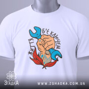 Футболка життя б`є ключем. Біла футболка складена, на ній видно принт з рукою, що тримає синій ключ, оточену червоними листям. Фон світлий, футболка зображена зверху.