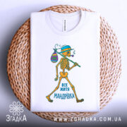 Футболка життя мандрівка, біла, зображена зверху, на фоні плетеного кошика, принт з малюнком скелета, який йде з мішком, текст "ВСЕ ЖИТТЯ МАНДРІВКА" внизу.