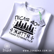 Футболка життя після 40, білого кольору, зображена зверху, на ній видно етикетку з зеленим шнурком, фон світлий, принт чорного кольору з написом 'ПІСЛЯ 40° ЖИТТЯ ТІЛЬКИ ПОЧИНАЄТЬСЯ' та пляшками.