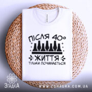 Футболка життя після 40, білого кольору, складена на плетеній кошику, фон світлий, принт чорного кольору з написом 'ПІСЛЯ 40° ЖИТТЯ ТІЛЬКИ ПОЧИНАЄТЬСЯ' та пляшками.
