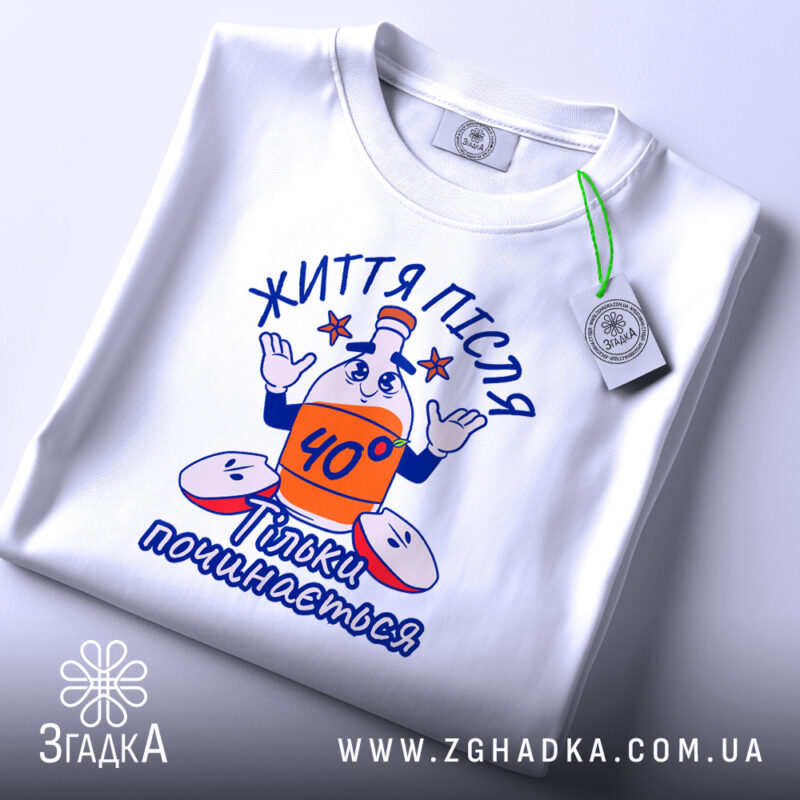 Футболка життя після 40, білого кольору, складена на столі, фон світлий, принт з персонажем, що тримає пляшку з написом '40°' і фразою 'Тільки починається', на етикетці видно логотип бренду.