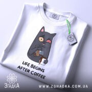 Футболка життя після кави, складена на поверхні, з видимим етикеткою, фон світлий, принт з котом, що п'є каву, з виразом обличчя, що показує емоції, текст під принтом чорного кольору.