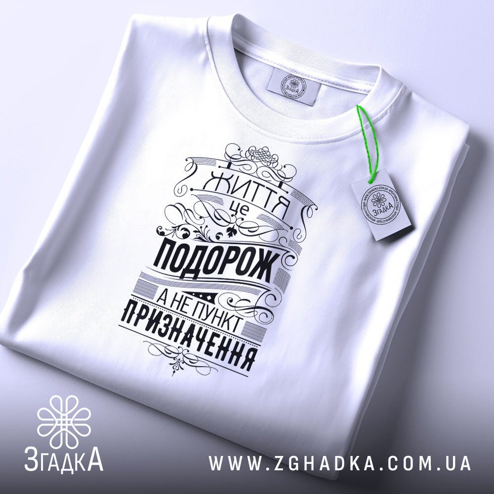 Футболка життя це подорож. Біла футболка, складена на поверхні, з видимим принтом чорного кольору. На футболці напис "Життя це подорож, а не пункт призначення" у стилі ретро, з етикеткою зеленого кольору, фон світло-сірий.