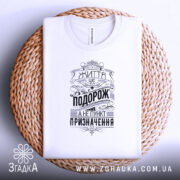 Футболка життя це подорож. Біла футболка, складена на плетеній основі, з видимим принтом чорного кольору. На футболці напис "Життя це подорож, а не пункт призначення" у стилі ретро, фон світло-сірий.