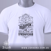 Футболка життя це подорож. Біла футболка, складена на поверхні, з видимим принтом чорного кольору. На футболці напис "Життя це подорож, а не пункт призначення" у стилі ретро, з етикеткою зеленого кольору, фон світло-сірий.