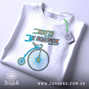 Футболка життя це подорож, біла футболка, складена на поверхні, з принтом велосипеда, видно етикетку з зеленим шнурком, фон світлий, принт яскравий.