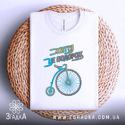 Футболка життя це подорож, біла футболка, складена на плетеній кошику, з принтом велосипеда, фон світлий, принт яскравий і контрастний.