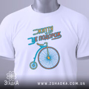 Футболка життя це подорож, біла футболка, складена на поверхні, з принтом велосипеда, видно етикетку з зеленим шнурком, фон світлий, принт яскравий.