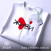 Футболка живи білого кольору, складена на плоскій поверхні, фон світло-сірий, принт з червоним серцем та чорним написом 'ЖИВИ', на комірці видна етикетка.