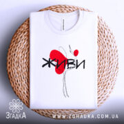 Футболка живи білого кольору, розташована на плоскій поверхні, фон світло-сірий, принт з червоним серцем та чорним написом 'ЖИВИ', футболка лежить на плетеній основі.