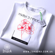 Футболка живи з мрією, білого кольору, складена на поверхні, фон світлий, принт з червоними квітами та написом, на футболці є етикетка.