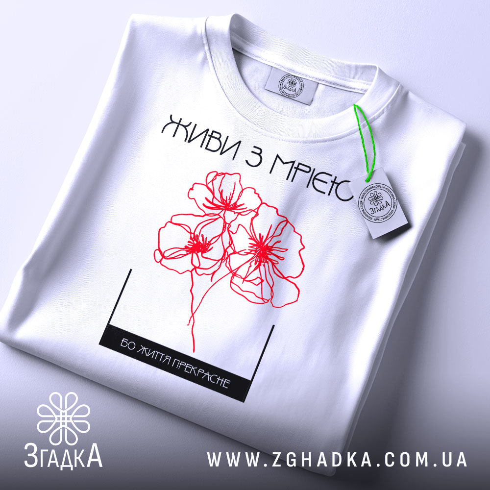Футболка живи з мрією, білого кольору, складена на поверхні, фон світлий, принт з червоними квітами та написом, на футболці є етикетка.