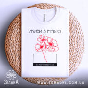 Футболка живи з мрією, білого кольору, складена на плетеній основі, фон світлий, принт з червоними квітами та написом, видима етикетка.