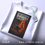 Футболка живи життя дрон, біла, складена на поверхні, фон світло-сірий, принт з дроном на фоні червоного неба та Кремлівської вежі, текст 'ЖИВИ ЖИТТЯ' у верхній частині принта, з етикеткою на комірці.