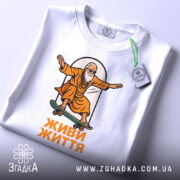 Футболка живи життя, біла, зображена в складеному вигляді, на фоні світло-сірого градієнту, принт з малюнком старця на скейтборді в оранжевому одязі, текст 'ЖИВИ ЖИТТЯ' під малюнком, на етикетці зелена стрічка.