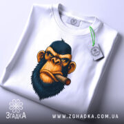 Футболка зла мавпа, біла, складена на столі, фон світло-сірий, принт з обличчям мавпи з сигарою, на футболці є етикетка з зеленим шнурком.