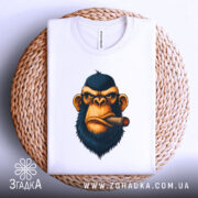 Футболка зла мавпа, біла, складена на кошику, фон світло-сірий, принт з обличчям мавпи з сигарою, деталі принта яскраві, футболка розташована на плетеній основі.