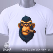 Футболка зла мавпа, біла, складена на столі, фон світло-сірий, принт з обличчям мавпи з сигарою, на футболці є етикетка з зеленим шнурком.