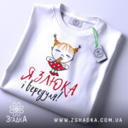 Футболка злюка вередуля, білого кольору, складена на поверхні, фон світло-сірий, принт з малюнком дівчинки з рудими волоссям у двох кісках, що тримає цукерку, текст червоним і чорним кольором, на комірці видна етикетка.