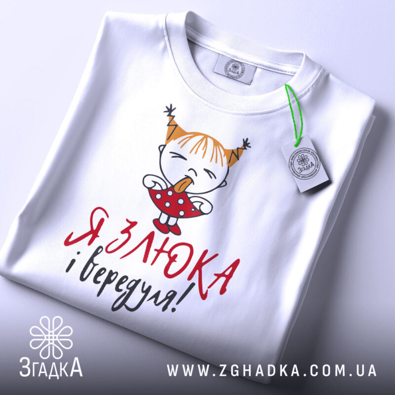 Футболка злюка вередуля, білого кольору, складена на поверхні, фон світло-сірий, принт з малюнком дівчинки з рудими волоссям у двох кісках, що тримає цукерку, текст червоним і чорним кольором, на комірці видна етикетка.