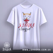 Футболка злюка вередуля, білого кольору, підвішена на вішалці, фон світло-сірий, принт з малюнком дівчинки з рудими волоссям у двох кісках, що тримає цукерку, текст червоним і чорним кольором.