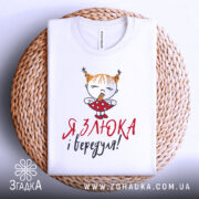 Футболка злюка вередуля, білого кольору, складена на плетеній кошику, фон білий, принт з малюнком дівчинки з рудими волоссям у двох кісках, що тримає цукерку, текст червоним і чорним кольором.