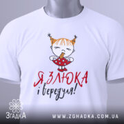 Футболка злюка вередуля, білого кольору, складена на поверхні, фон світло-сірий, принт з малюнком дівчинки з рудими волоссям у двох кісках, що тримає цукерку, текст червоним і чорним кольором, на комірці видна етикетка.