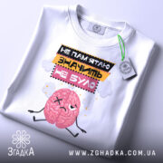 Футболка значить не було. Біла футболка, складена на поверхні, з принтом мозку, який виглядає сумно. Текст на футболці: 'НЕ ПАМ'ЯТЮ ЗНАЧИТЬ НЕ БУЛО'. На футболці є етикетка з зеленим шнурком. Фон світлий, з легким відблиском.