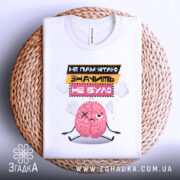 Футболка значить не було. Біла футболка, складена на плетеній основі, з принтом мозку, що виглядає сумно. Текст на футболці: 'НЕ ПАМ'ЯТЮ ЗНАЧИТЬ НЕ БУЛО'. Фон світлий, з текстурою плетеного матеріалу.