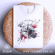 Футболка знайди свій шлях, біла, складена на плетеній кошику, фон білий, принт з компасом та червоними плямами фарби.