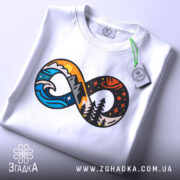 Футболка знак нескінченності, біла, складена на поверхні, фон світло-сірий, принт з елементами гір, хвиль та лісу в яскравих кольорах, з зеленим ярликом.